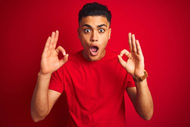 İzole edilmiş kırmızı arka plan üzerinde duran Brezilyalı genç adam şaşırmış ve şaşırmış görünüyor. Tamam sembolünü parmaklarıyla yapıyor. Çılgın ifade