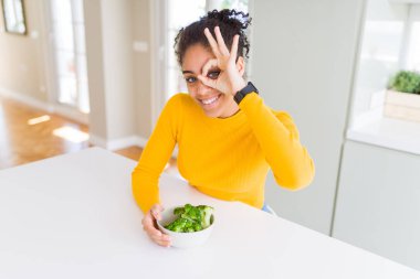 Genç Afro-Amerikalı kız sıcak yeşil brokoli yiyor, gülümseyen bir gülümseme ile tamam işaret ediyor, parmaklarının arasından bakıyor.