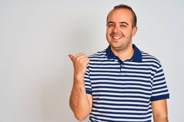 Sıradan çizgili polo giyen genç adam izole edilmiş beyaz arka planda durup gülümsüyor ve baş parmağıyla yan tarafa bakıyor..