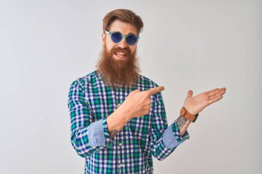 İzole edilmiş beyaz arka plan üzerinde gündelik gömlek ve güneş gözlüğü takan genç kızıl saçlı adam kameraya hayret ediyor ve gülümsüyor..