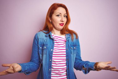 Güzel kızıl saçlı kadın kot gömlek ve çizgili tişört giyerek izole edilmiş pembe arka plan üzerinde habersiz ve açık kollarla kafası karışmış, hiçbir fikri yok..