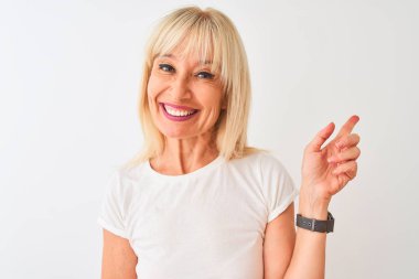 İzole edilmiş beyaz arka planda duran sıradan tişört giyen orta yaşlı bir kadın mutlu bir şekilde elini ve parmağını kenarı işaret ediyor.