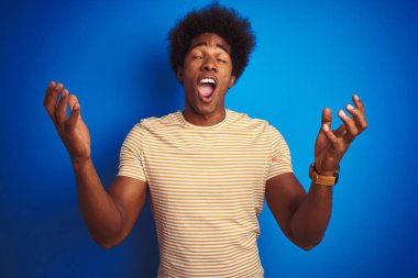 İzole edilmiş mavi arka plan üzerine çizgili sarı t-shirt giyen Amerikalı bir adam başarı için deli ve çılgınlığını kollarını kaldırarak ve gözlerini kapayarak heyecanla bağırarak kutluyor. Kazanan konsept