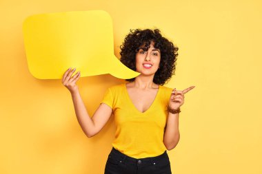 Kıvırcık saçlı genç Arap kadın izole edilmiş sarı arka planda konuşma baloncuğu tutuyor. Elleri ve parmakları kenarı işaret ederek çok mutlu.