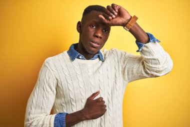 İzole edilmiş sarı arka planda kot pantolon ve beyaz kazak giyen Afro-Amerikan bir adam. Hastalık, ateş, grip ve soğuk algınlığı için alnına dokunuyor.