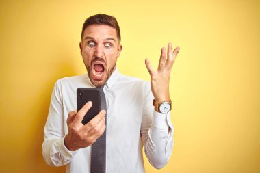 Genç yakışıklı iş adamı, sarı arka plan üzerinde akıllı telefon kullanıyor. Çok mutlu ve heyecanlı, kazanan yüz ifadesi zaferi büyük bir gülümsemeyle kutluyor ve ellerini kaldırıyor.