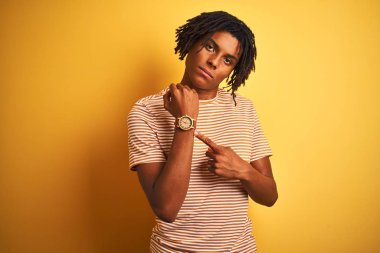 Sarı arka plan üzerinde duran çizgili t-shirt giyen Afro bir adam. Aceleyle saati, sabırsızlığı ve rahat bir ifadeyle kameraya bakıyor.