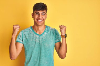 Yeşil t-shirt giyen genç Hintli adam, izole edilmiş sarı arka planda duruyor. Şaşırmış ve şaşırmış bir şekilde kollarını kaldırıp, gözlerini açarak başarıyı kutluyor. Kazanan konsept.