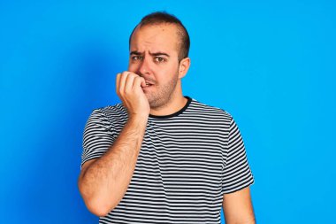 İzole edilmiş mavi arka planda duran çizgili denizci tişörtü giyen genç adam elleri ağız yiyen tırnaklarla gergin ve stresli görünüyor. Endişe sorunu.