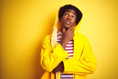 Kapüşonlu bir yağmurluk giyen afro saçlı Afrikalı bir adam izole edilmiş sarı arka planda duruyor, elleri çenesinde soru sorarken, düşünceli bir ifade takınıyor. Düşünceli bir yüzle gülümsüyor. Şüphe kavramı.