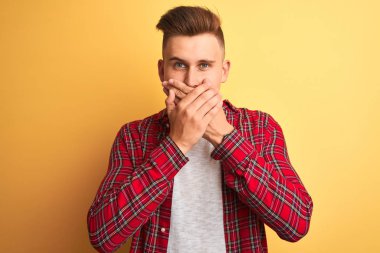 Izole sarı arka plan üzerinde duran rahat gömlek giyen genç yakışıklı adam hata için elleri ile ağız kapsayan şok. Gizli kavram.