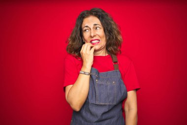 Kırmızı izole arka planda önlük giyen orta yaşlı bir kadın elleri ağız yiyen tırnaklarla gergin ve gergin görünüyor. Endişe sorunu.