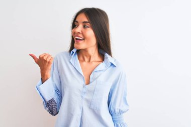 Genç, güzel bir kadın, mavi şık bir gömlek giyiyor. İzole edilmiş beyaz arka planda duruyor. Gülümsüyor, gülümsüyor ve başparmağıyla yan tarafa bakıyor..