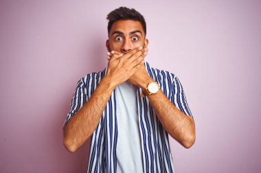 İzole edilmiş pembe arka plan üzerine yaz çizgili tişört giyen genç ve yakışıklı Hintli adam yanlışlıkla ağızlarını kapatarak şok etti. Gizli konsept.