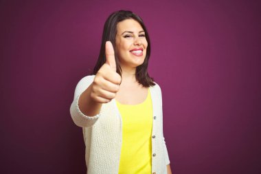 Genç, güzel, esmer bir kadın. Mor izole bir arka plan üzerinde bir ceket. Elle mutlu bir jest yapıyor. Kameraya bakıp başarıyı gösteren ifade onaylanıyor.