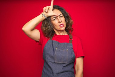 Kırmızı izole arka planda önlük giyen orta yaşlı bir kadın. Alnında parmakları olan insanlarla alay ediyor. Ezik hareketler yapıyor, alay ediyor..