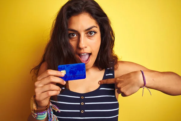 Young beautiful woman holding credit card standing over isolated yellow background with surprise face pointing finger to himself