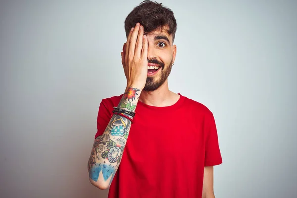 Young man with tattoo wearing red t-shirt standing over isolated white background covering one eye with hand, confident smile on face and surprise emotion.