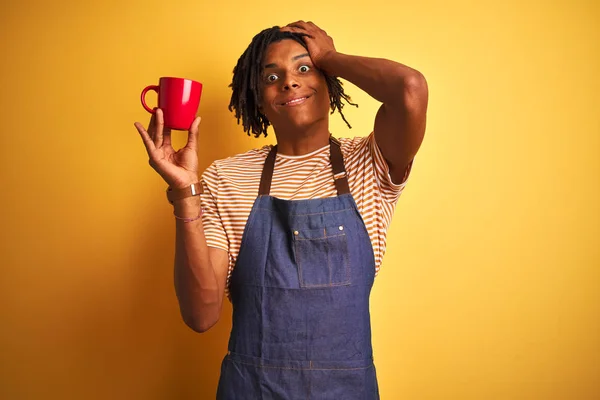 Afro barista man with dreadlocks drinking cup of coffee over isolated yellow background stressed with hand on head, shocked with shame and surprise face, angry and frustrated. Fear and upset for mistake.