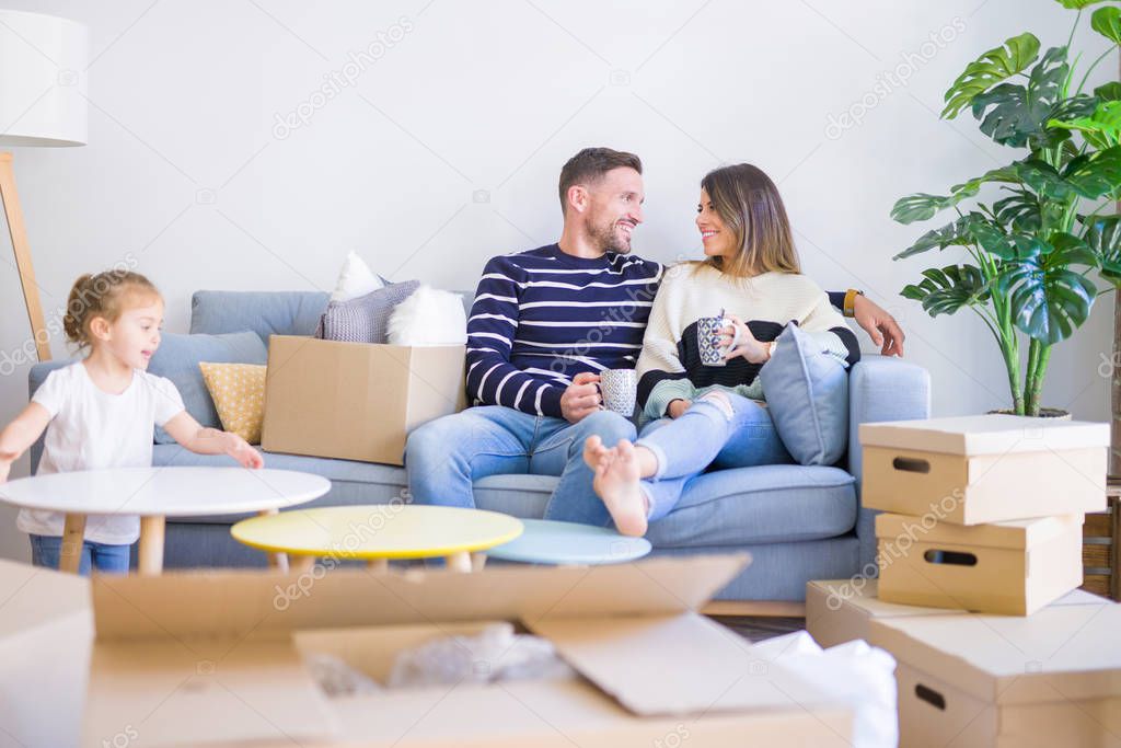 Beautiful family, parents sitting on the sofa drinking coffee looking his kid playing at new home around cardboard boxes