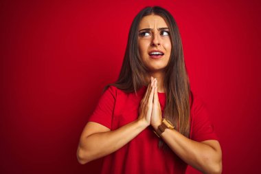 İzole edilmiş kırmızı arka plan üzerinde duran tişört giyen genç ve güzel bir kadın elleri bağlı yalvarıyor ve dua ediyor. Yüzünde umut ifadesi var. Çok duygusal ve endişeli. Af diliyor. Din kavramı.
