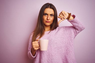Genç güzel kadın kızgın yüz ile izole pembe arka plan üzerinde duran kahve fincan içme, başparmak aşağı, ret kavramı ile sevmediğimi gösteren negatif işareti
