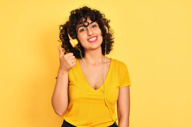 Kıvırcık saçlı, müzik dinleyen Arap bir kadın izole edilmiş sarı arka planda kulaklık takıyor ve elleriyle mutlu bir jest yapıyor. Kameraya bakıp başarıyı gösteren ifade onaylanıyor.