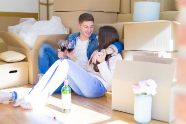 Karton kutular etrafında yeni evin katında oturan genç çift, şarap bir bardak içme kutluyor ve yeni daire için mutlu gülümseyen