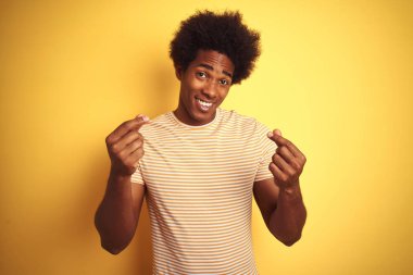 Afro saçlı, çizgili tişörtlü bir Amerikalı izole edilmiş sarı arka planda duruyor. Elleriyle para hareketi yapıyor. Maaş ödemesi istiyor.