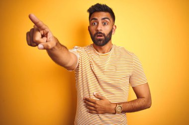 İzole edilmiş sarı arkaplanda duran tişörtlü genç Hintli bir adam. Parmakları önde, ağzı açık, şaşırmış, ön tarafta bir şey var.