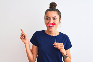 Genç, güzel bir kadın, izole edilmiş beyaz bir arka planda parti bıyığı tutuyor. Çok mutlu bir şekilde elini ve parmağını kenarı gösteriyor.