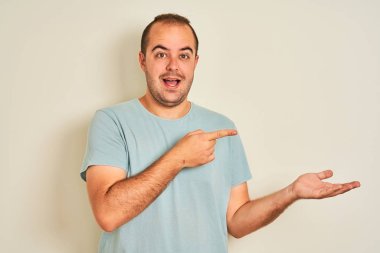 İzole edilmiş beyaz arka plan üzerinde duran mavi günlük tişört giyen genç adam hayrete düşmüş ve kameraya gülümserken eliyle ve parmağıyla işaret ediyordu..