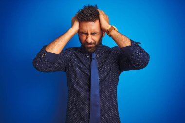 Genç Hintli iş adamı zarif gömlek ve kravat giyerek izole edilmiş mavi arka planda dikiliyor. Ağrı ve migrenden dolayı baş ağrısı ve stresten muzdarip. Ellerini başının üstüne koy..