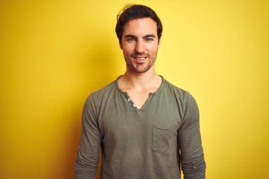 Yüzünde mutlu ve havalı bir gülümsemeyle izole edilmiş sarı arka plan üzerinde duran sıradan tişört giyen genç yakışıklı bir adam. Şanslı kişi..