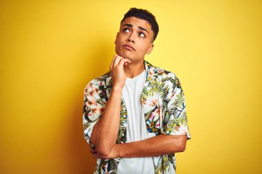 Tatilde olan genç Brezilyalı bir adam, çiçekli bir tişört giyiyor, izole edilmiş sarı arka planda, elleri çenesinde soru sorarken, düşünceli bir ifade takınıyor. Düşünceli bir yüzle gülümsüyor. Şüphe kavramı.