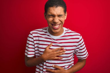İzole edilmiş kırmızı arka plan üzerinde çizgili tişört giyen genç, yakışıklı Arap adam gülmekten çatlıyor çünkü elleri vücutta komik bir şaka var..