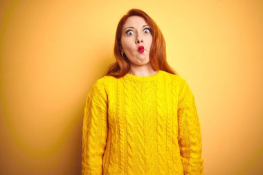 Kışlık süveter giyen güzel bir kızıl kadın izole edilmiş sarı arka planda duruyor dudaklı balık suratı yapıyor, çılgınca ve komik hareketler yapıyor. Komik ifade.