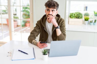 Genç bir öğrenci akıllı telefon kullanıyor ve laptopla çalışıyor. Ciddi bir yüz ifadesi kullanıyor. Soru hakkında düşünüyor, kafası karışık bir fikir.