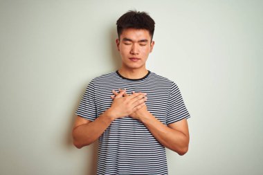 Asyalı, çizgili tişört giyen genç bir Çinli izole edilmiş beyaz arka planda duruyor elleri göğsünde, gözleri kapalı ve yüzünde minnet dolu bir jest var. Sağlık kavramı.