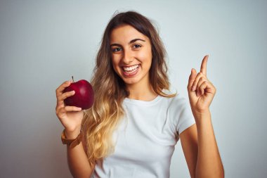 Gri arka planda kırmızı elma yiyen genç ve güzel bir kadın. Elleri ve parmakları kenarda mutlu bir şekilde işaret ediyor.