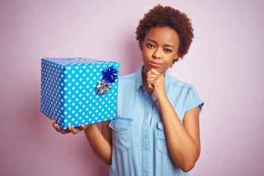 Afrika kökenli Amerikalı bir kadın pembe izole edilmiş arka planda doğum günü hediyesi tutuyor. Ciddi bir yüz ifadesi var. Soru sormayı düşünüyor.