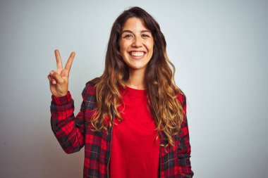 Kırmızı tişört ve ceket giyen genç ve güzel bir kadın beyaz izole edilmiş arka plan üzerinde duruyor ve parmaklarıyla iki numarayı gösterirken kendinden emin ve mutlu bir şekilde gülümsüyor..