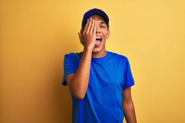 Yakışıklı Arap teslimatçı, izole edilmiş sarı arka planda duruyor. Bir gözünü eliyle kapatıyor. Yüzünde kendine güvenen bir gülümseme ve sürpriz bir duygu var..