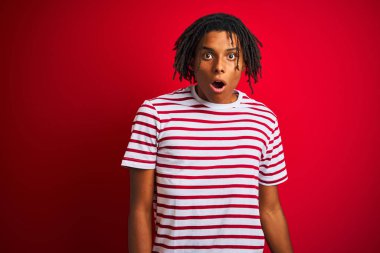 Rastalı tişörtlü genç afro adam izole edilmiş kırmızı arka planda dikiliyor. Şaşırmış ve şaşırmış bir ifade, korku ve heyecanlı bir yüz ifadesi var..
