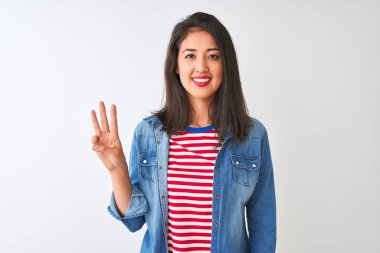 İzole edilmiş beyaz arka plan üzerinde çizgili tişört ve kot giyen genç Çinli kadın üç numarayı gösterirken kendinden emin ve mutlu bir şekilde gülümsüyor..