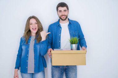 Genç çift ellerinde karton kutuyla beyaz izole bir zemin üzerinde yeni bir eve taşınıyor ve mutlu bir gülümsemeyle baş parmağıyla işaret ediyor.