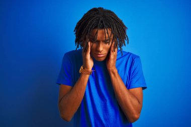 Rastalı tişörtlü Afro-Amerikan bir adam izole edilmiş mavi arka planda dikiliyor ve baş ağrısı çekiyor çünkü stres var. Migren ağrısı..