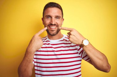 Sarı izole edilmiş arka plan üzerine gündelik kırmızı çizgili tişört giyen, neşeli gülümseyen ve parmaklarını ve ağzını gösteren genç yakışıklı bir adam. Diş sağlığı konsepti.