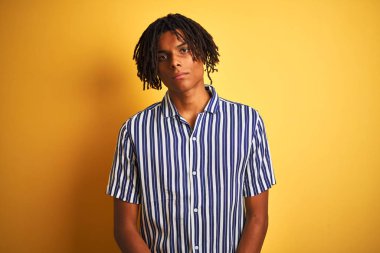 Rasta desenli tişörtlü Afro adam izole edilmiş sarı arka plan üzerinde rahatlamış ve yüzünde ciddi bir ifade vardı. Basit ve doğal bir şekilde kameraya bakıyorsun..