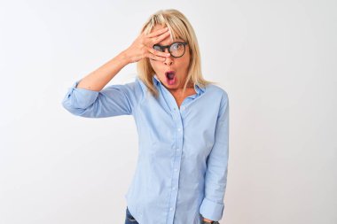 Orta yaşlı iş kadını zarif bir gömlek ve gözlük takıyor, izole edilmiş beyaz bir arkaplanı var. Şok olmuş bir şekilde yüzünü ve gözlerini kapatıyor, utanmış bir ifadeyle parmaklarından bakıyor..